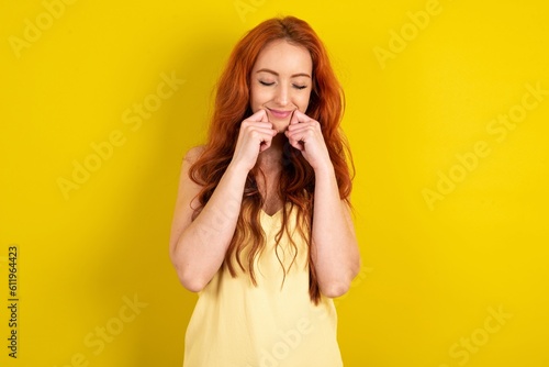 Pleased young redhead woman wearing yellow t-shirt over yellow wall with closed eyes keeps hands near cheeks and smiles tenderly imagines something very pleasant