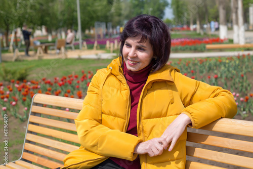 An adult happy woman with a smile on her face in a bright yellow jacket is resting on a bench in the park..