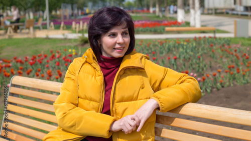 An adult happy woman with a smile face in a bright yellow jacket is resting on a bench in the park..