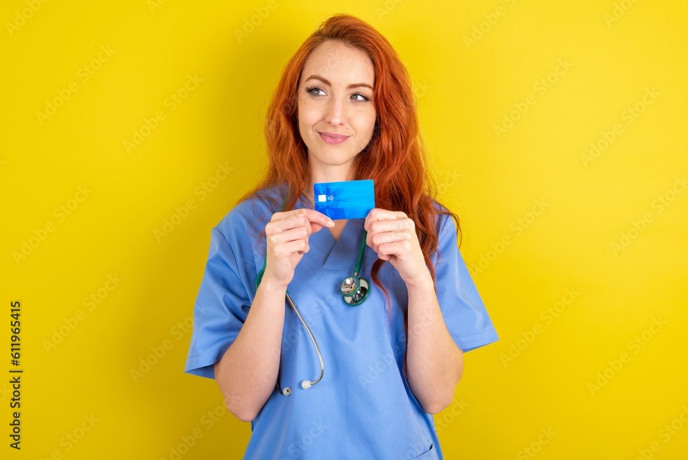 Photo of cheerful redhead doctors woman wearing blue uniform over yellow wall hold debit card look empty space Photo of cheerful redhead doctors woman wearing blue uniform over yellow wall hold debit card look empty space