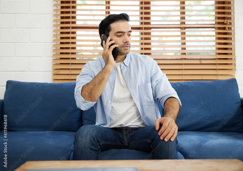 Fototapeta premium young handsome man sitting on sofa and talking by smartphone at home