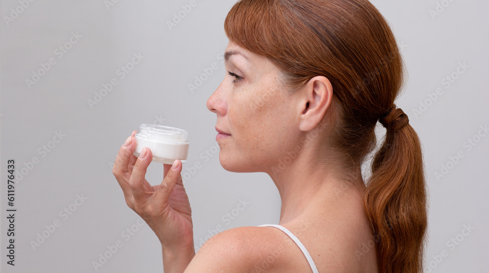Back of cropped caucasian middle aged woman face with freckles holding opened jar with skin cream in hand on white background Back of cropped caucasian middle aged woman face with freckles holding opened jar with skin cream in hand on white background
