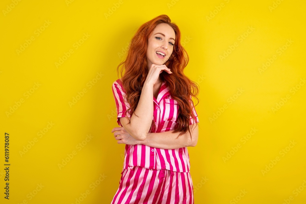 Optimistic Young red haired woman wearing pink pyjama over yellow studio background keeps hands partly crossed and hand under chin, looks at camera with pleasure. Optimistic Young red haired woman wearing pink pyjama over yellow studio background keeps hands partly crossed and hand under chin, looks at camera with pleasure.
