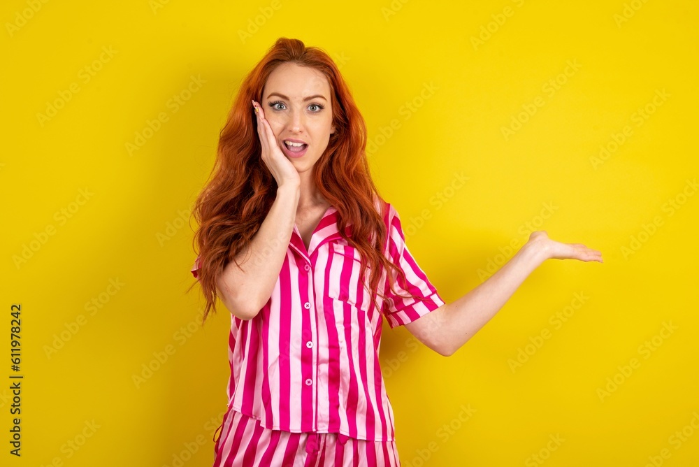 Crazy Young red haired woman wearing pink pyjama over yellow studio background advising discount prices hold open palm new product