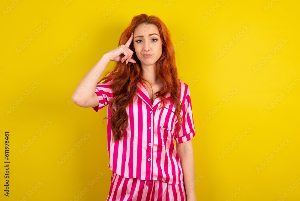 Young red haired woman wearing pink pyjama over yellow studio background tries to memorize something, keeps fore finger on temple, reminds information for exam, Young red haired woman wearing pink pyjama over yellow studio background tries to memorize something, keeps fore finger on temple, reminds information for exam,