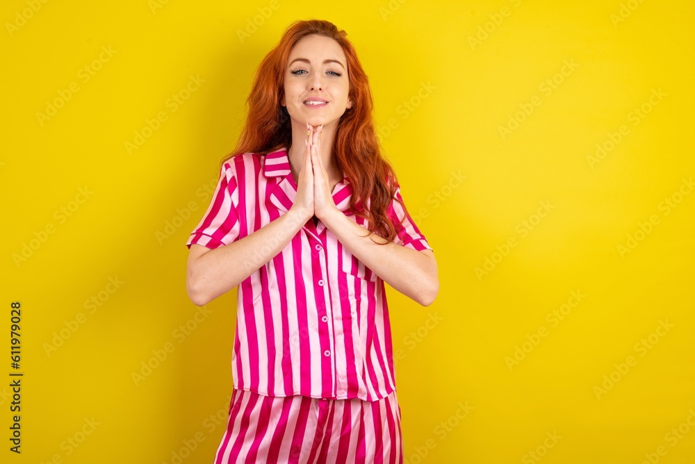Young red haired woman wearing pink pyjama over yellow studio background praying with hands together asking for forgiveness smiling confident. Young red haired woman wearing pink pyjama over yellow studio background praying with hands together asking for forgiveness smiling confident.