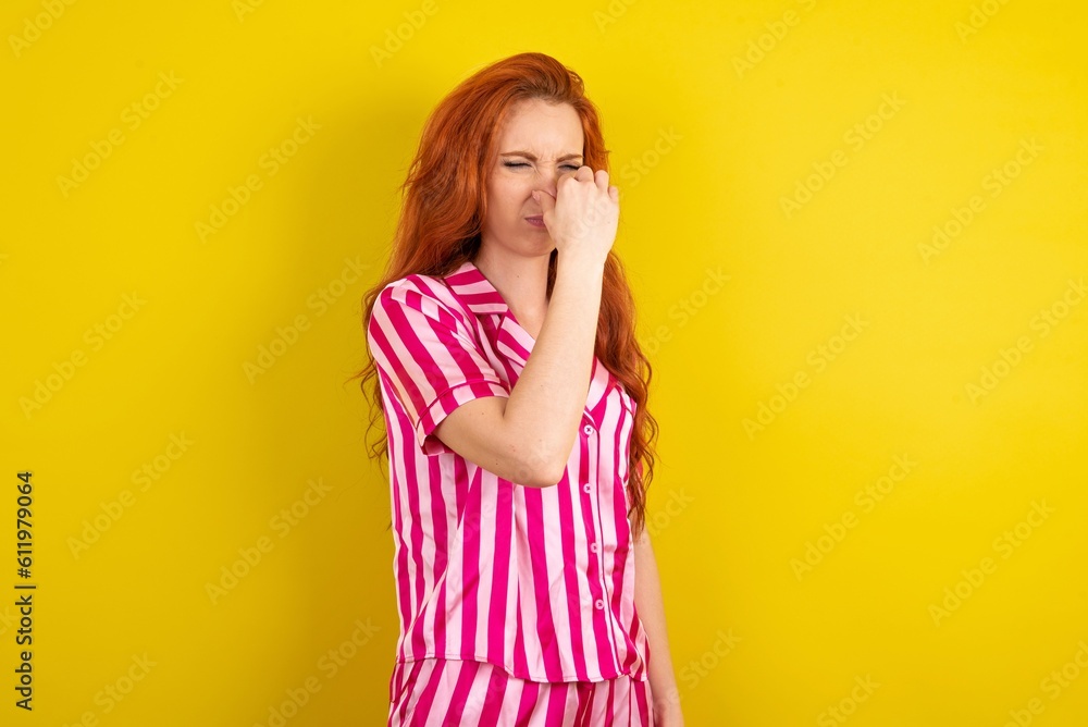 Young red haired woman wearing pink pyjama over yellow studio background smelling something stinky and disgusting, intolerable smell, holding breath with fingers on nose. Bad smell Young red haired woman wearing pink pyjama over yellow studio background smelling something stinky and disgusting, intolerable smell, holding breath with fingers on nose. Bad smell