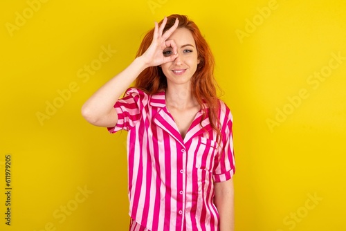 Young red haired woman wearing pink pyjama over yellow studio background doing ok gesture with hand smiling, eye looking through fingers with happy face.