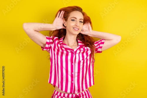 Young red haired woman wearing pink pyjama over yellow studio background Trying to hear both hands on ear gesture, curious for gossip. Hearing problem, deaf