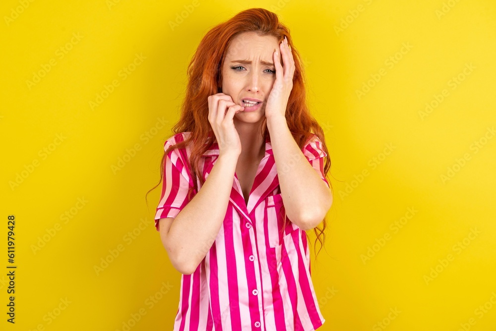 Doleful desperate crying Young red haired woman wearing pink pyjama over yellow studio background, looks stressfully, frowns face, feels lonely and anxious Doleful desperate crying Young red haired woman wearing pink pyjama over yellow studio background, looks stressfully, frowns face, feels lonely and anxious