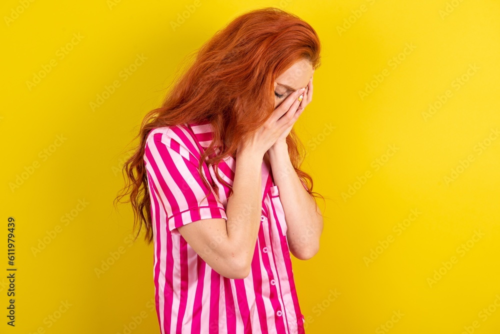 Sad Young red haired woman wearing pink pyjama over yellow studio background crying covering her face with her hands. Sad Young red haired woman wearing pink pyjama over yellow studio background crying covering her face with her hands.