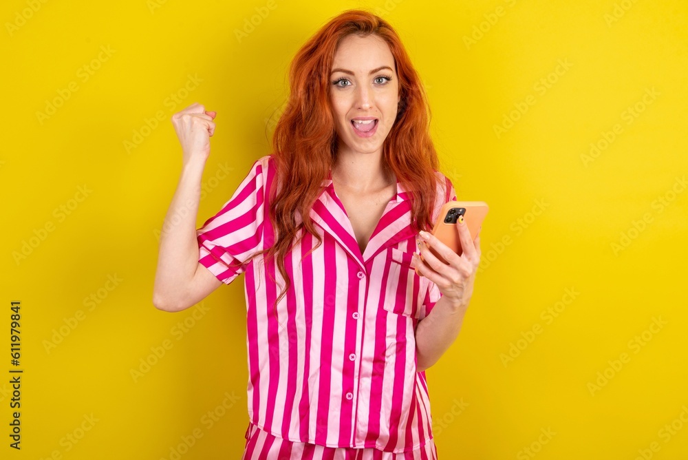 Portrait of Young red haired woman wearing pink pyjama over yellow studio background holding in hands cell celebrating Portrait of Young red haired woman wearing pink pyjama over yellow studio background holding in hands cell celebrating
