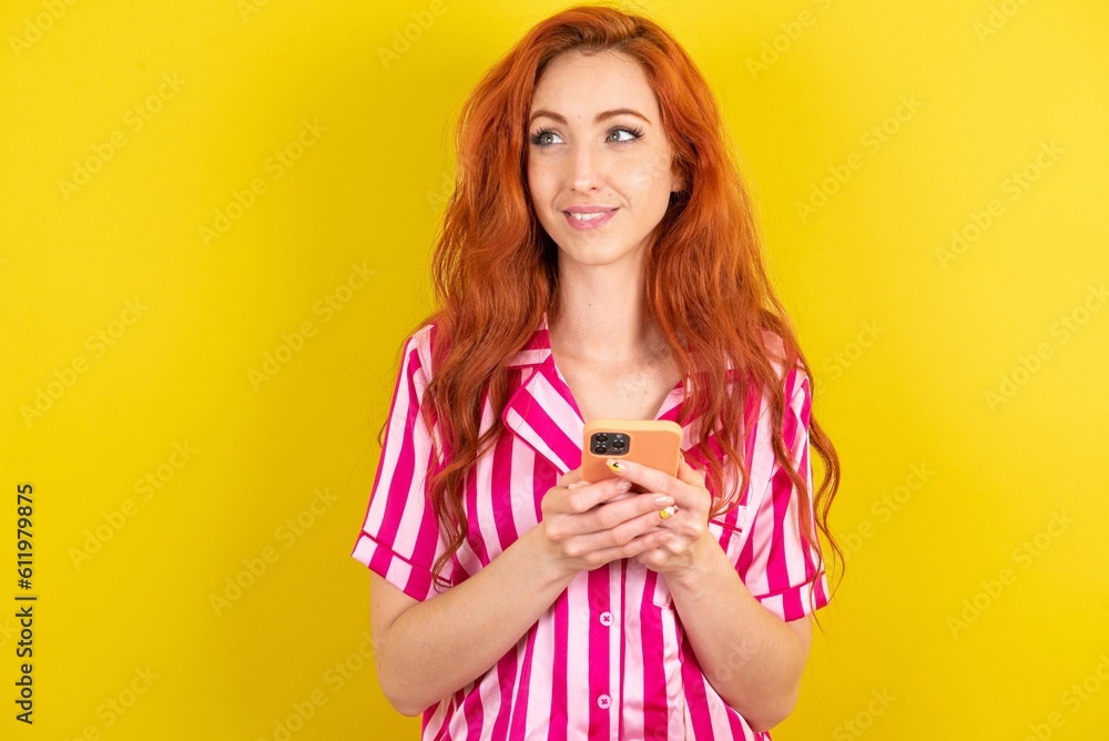 Young red haired woman wearing pink pyjama over yellow studio background holding in hands mobile phone and looking aside with dreamy look. Young red haired woman wearing pink pyjama over yellow studio background holding in hands mobile phone and looking aside with dreamy look.