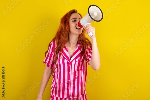 Young red haired woman wearing pink pyjama over yellow studio background Through Megaphone with Available Copy Space