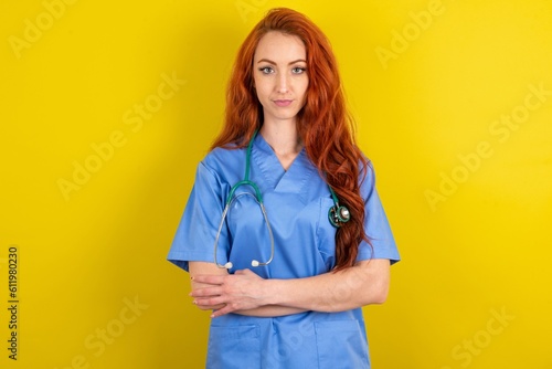 Self confident serious calm young red-haired doctor woman over yellow studio background stands with arms folded. Shows professional vibe stands in assertive pose.