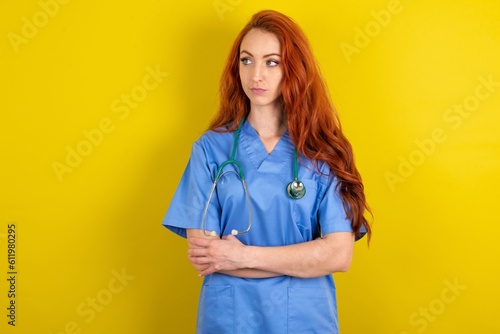 Charming thoughtful young red-haired doctor woman over yellow studio background stands with arms folded concentrated somewhere with pensive expression thinks what to do