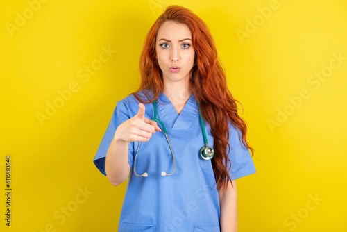 Shocked young red-haired doctor woman over yellow studio background  points at you with stunned expression