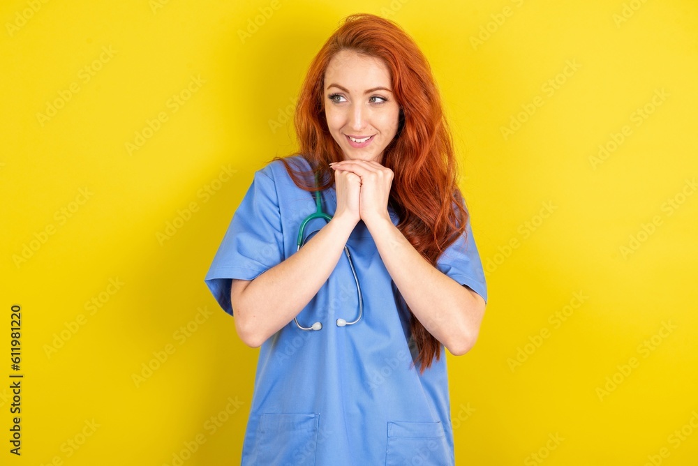 Happy young red-haired doctor woman over yellow studio background anticipates something awesome happen, looks happily aside, keeps hands together near face, has glad expression. Happy young red-haired doctor woman over yellow studio background anticipates something awesome happen, looks happily aside, keeps hands together near face, has glad expression.