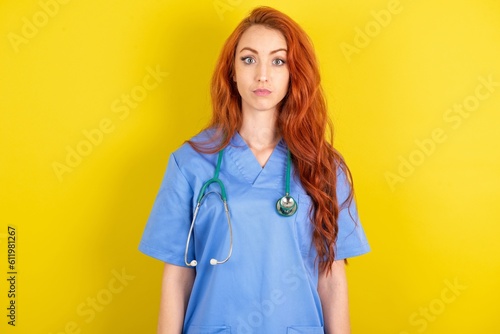 Stunned young red-haired doctor woman over yellow studio background stares reacts on shocking news. Astonished girl holds breath