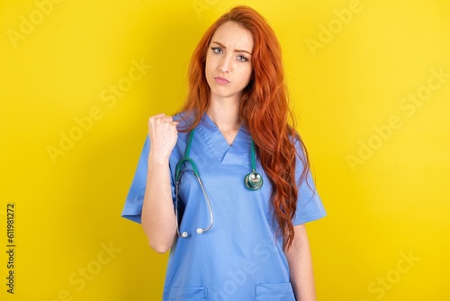 young red-haired doctor woman over yellow studio background shows fist has annoyed face expression going to revenge or threaten someone makes serious look. I will show you who is boss