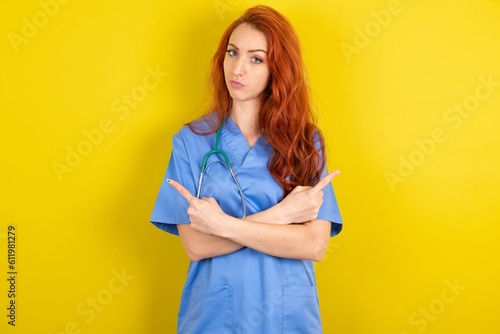 Serious young red-haired doctor woman over yellow studio background crosses hands and points at different sides hesitates between two items. Hard decision concept
