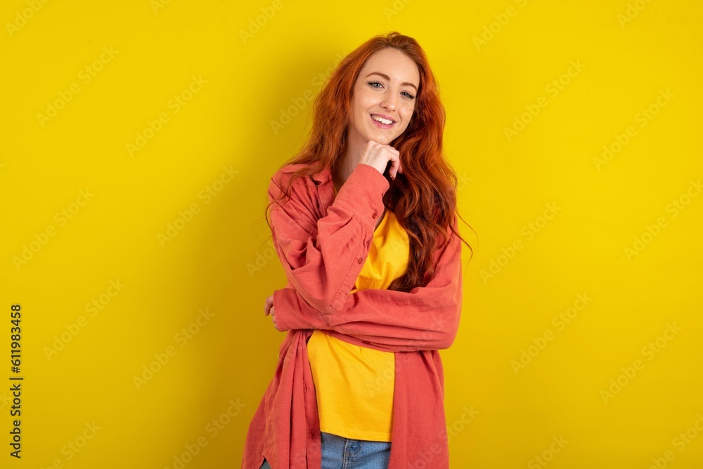 Optimistic young beautiful red haired woman over yellow studio background keeps hands partly crossed and hand under chin, looks at camera with pleasure. Optimistic young beautiful red haired woman over yellow studio background keeps hands partly crossed and hand under chin, looks at camera with pleasure.