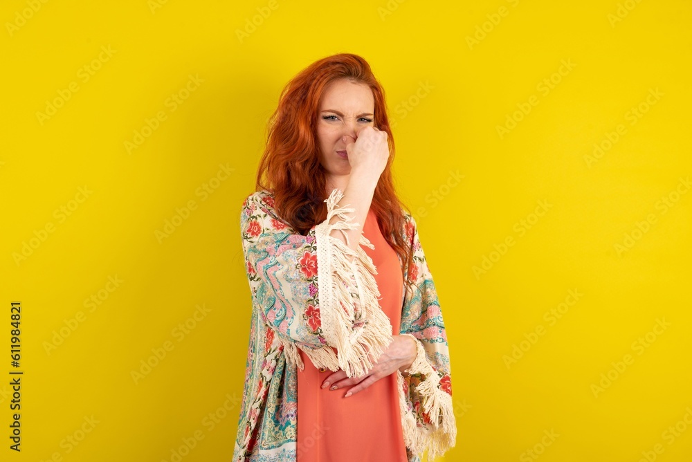 young beautiful red haired woman smelling something stinky and disgusting, intolerable smell, holding breath with fingers on nose. Bad smell