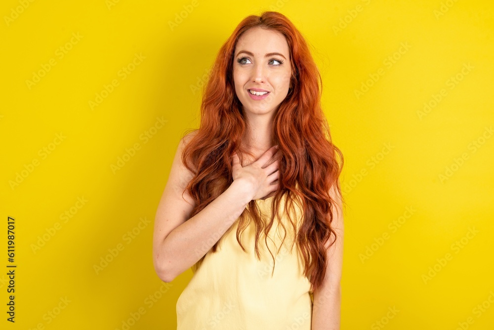 Joyful young beautiful red haired woman wearing yellow blouse over yellow studio background expresses positive emotions recalls something funny keeps hand on chest and giggles happily. Joyful young beautiful red haired woman wearing yellow blouse over yellow studio background expresses positive emotions recalls something funny keeps hand on chest and giggles happily.