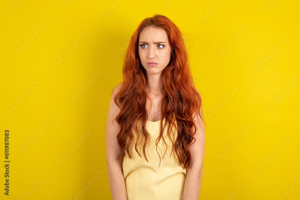 Fototapeta premium young beautiful red haired woman wearing yellow blouse over yellow studio background stares aside with wondered expression has speechless expression. Embarrassed model looks in surprise