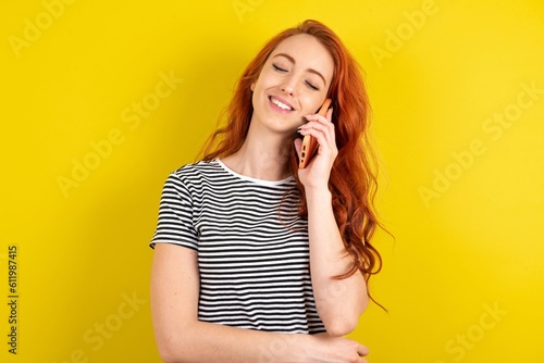 Funny young beautiful red haired woman wearing striped t shirt over yellow studio background laughs happily, has phone conversation, being amused by friend, closes eyes.