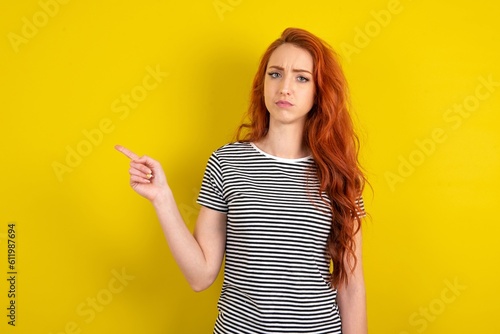 Serious young beautiful red haired woman wearing striped t shirt over yellow studio back smirks face points away on copy space shows something unpleasant. Look at this advertisement. Big price concept