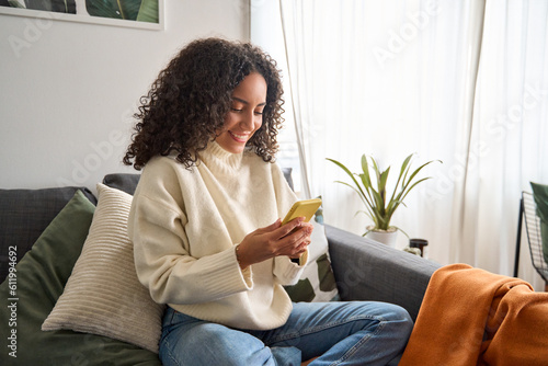 Fototapet Happy young latin woman sitting on sofa holding mobile phone using cellphone technology doing ecommerce shopping, buying online, texting messages relaxing on couch in cozy living room at home