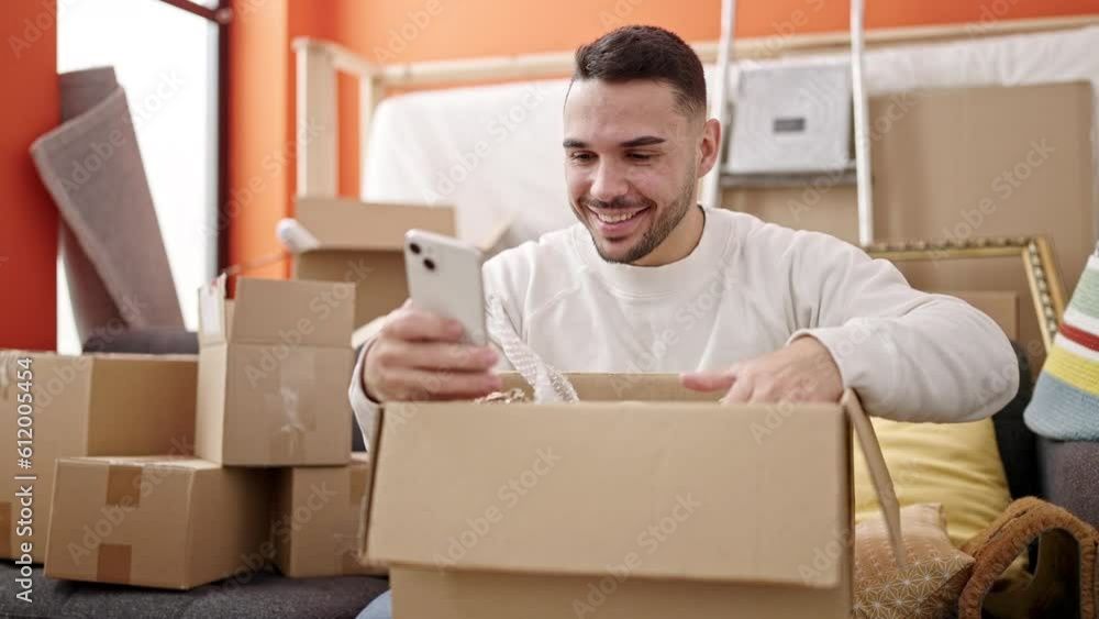 Young hispanic man unpacking cardboard box using smartphone at new home Young hispanic man unpacking cardboard box using smartphone at new home