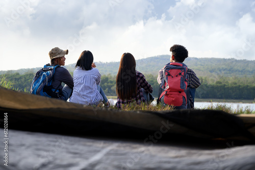 Asian young people groups sit and look at mountain lake views and relax near camping tents in the natural park. Enjoying freedom lifestyle and relaxation in summer with friendship concept.