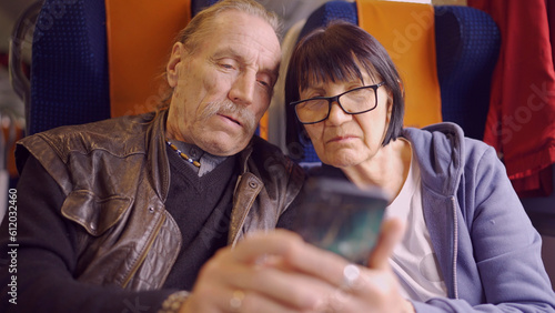 Elderly couple travel by train, the senior is holding a mobile phone in her hand, both are looking at the smartphone and talking with each other