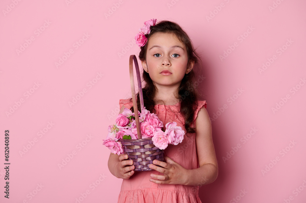 Studio portrait of a Caucasian noble cute little girl with a curly hair and beautiful blue eyes, dressed in a pink dress, holding a basket of roses, looking insightfully at camera over pink background