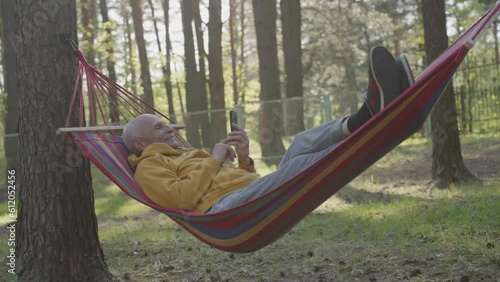 Elderly man with smile looks photos in mobile phone lying on colorful hammock attached to coniferous trees in city park. Recreation for old male person photo