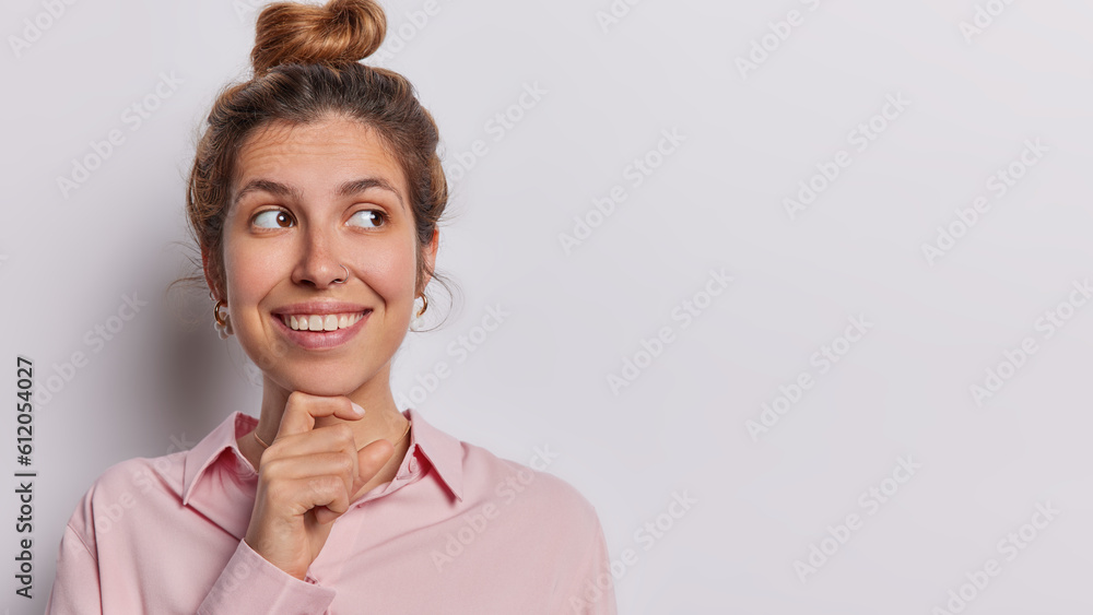 Studio shot of beautiful woman smiles toothily has gentle expression smiles tenderly keeps hand under chin concentrated wears shirt isolated on white background copy space for your advertising content Studio shot of beautiful woman smiles toothily has gentle expression smiles tenderly keeps hand under chin concentrated wears shirt isolated on white background copy space for your advertising content