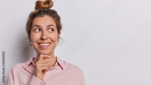Studio shot of beautiful woman smiles toothily has gentle expression smiles tenderly keeps hand under chin concentrated wears shirt isolated on white background copy space for your advertising content
