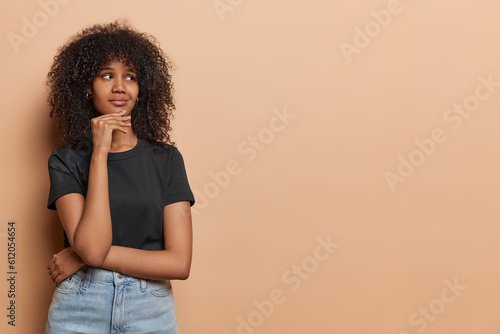 Horizontal shot of curly haired woman captured in moment of deep contemplation gazes into distance evoking sense of curiosity and wonder keeps hand on chin wears black t shirt jeans isolated on beige