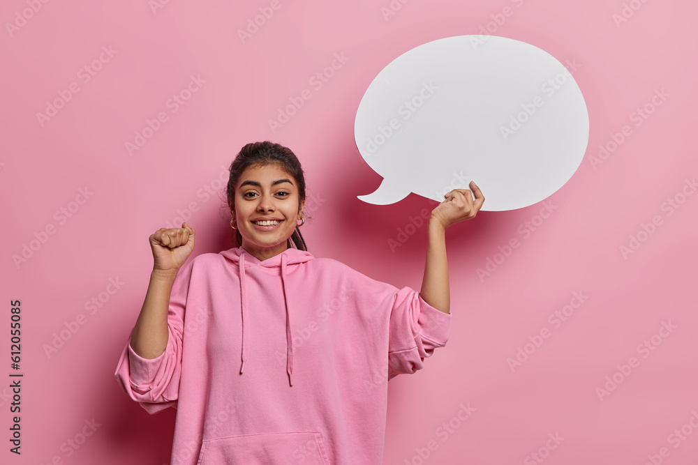 Cheerful Indian woman clenches fist and holds white blank speech bubble for writing your imaginative advertising content smiles broadly dressed in casual hoodie isolated over pink background Cheerful Indian woman clenches fist and holds white blank speech bubble for writing your imaginative advertising content smiles broadly dressed in casual hoodie isolated over pink background