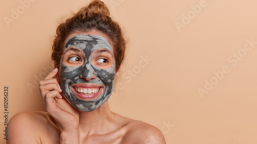 Horizontal shot of happy dark haired woman gracefully applies clay mask on her face promoting benefits of natural skincare gives skincare tutorials looks happily aside isolated over beige background
