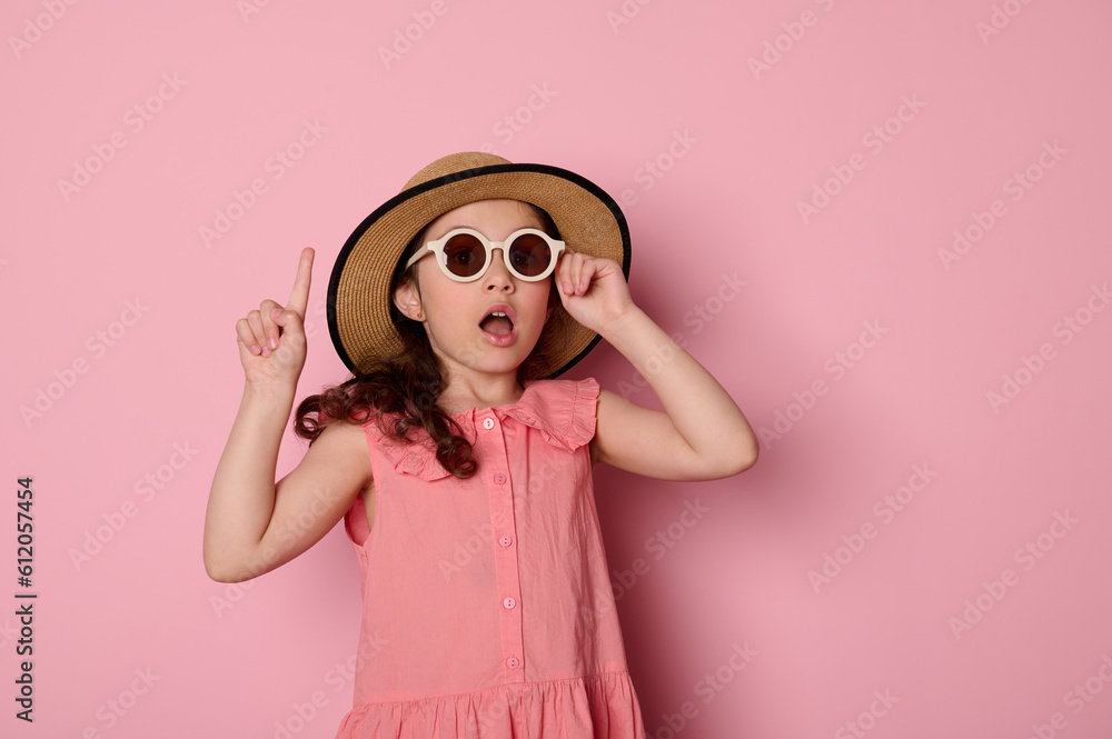 Amazed adorable child girl wearing sunglasses, pink dress and straw hat, pointing forefinger aside a copy space for your advertising text, on isolated pink background. Summer camp. Holidays concept Amazed adorable child girl wearing sunglasses, pink dress and straw hat, pointing forefinger aside a copy space for your advertising text, on isolated pink background. Summer camp. Holidays concept