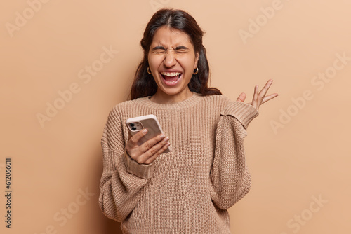 Woman with blonde curly hair exclaims loudly her raised palm capturing intensity of moment with mobile phone in hand she receives messages from both her boyfriend and friends dressed in knitted jumper