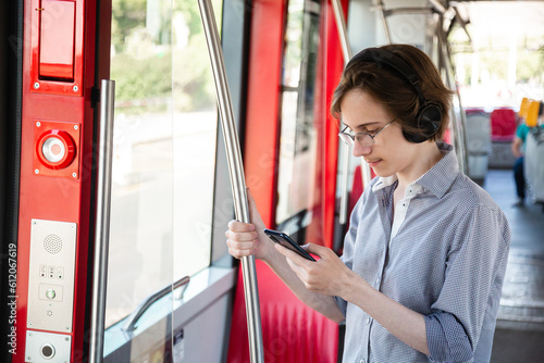 Tennager boy in headphones in tram holding mobile phone in his hand. Guy stays near door, looks with interest at screen of smartphone, playing mobile game online on smartphone connected to public wifi photo