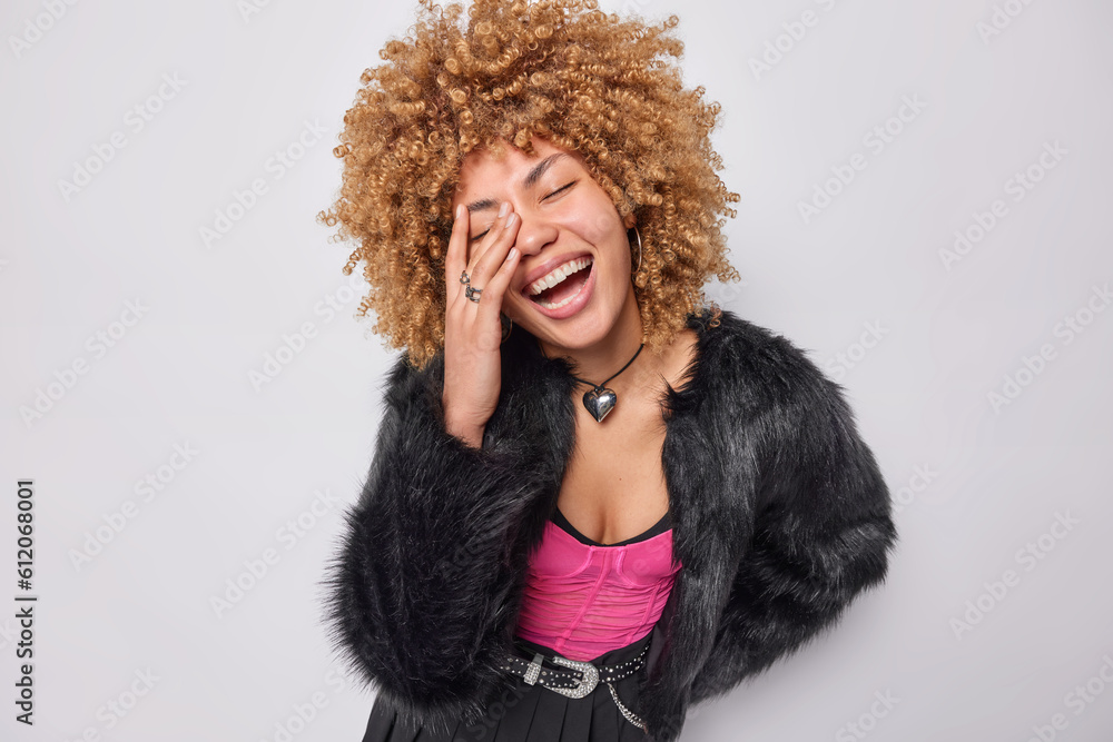 Joyful woman with curly hair makes face palm smiles broadly keeps eyes closed dressed in winter fur coat and pleated skirt isolated over white background. People positive emotions and fashion concept Joyful woman with curly hair makes face palm smiles broadly keeps eyes closed dressed in winter fur coat and pleated skirt isolated over white background. People positive emotions and fashion concept