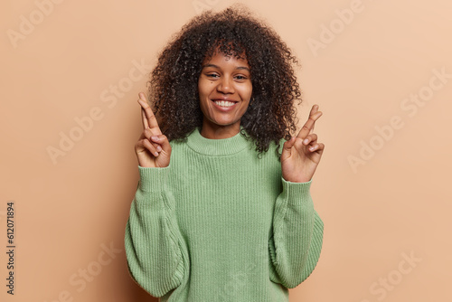 Pretty curly African woman stands with crossed fingers warm smile emanating strong belief in positive outcomes her faith in good fortune reflects spirit of hope resilience wears casual knitted jumper © wayhome.studio 