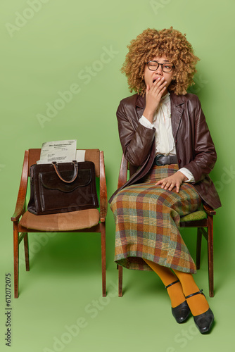 Retro woman sits on chair with tired expression yawns and covers mouth exuding timeless elegance with vintage attire and classic hairstyle poses on chair waits for something isolated over green wall