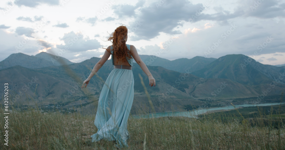 Beautiful carefree girl with red hair wearing white dress standing against the wind, looking at mountains and raising her hands - freedom, adventure, inspiration 