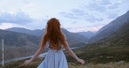 Carefree girl with red hair raising her hands up to the wind, embracing freedom and tranquility, enjoying scenic view of mountains 